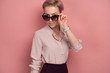 © bestsenny - A girl with short pink hair in a blouse and skirt stands on a pink background, looking over sunglasses.