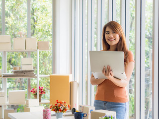 Wall Mural - Portrait of a beautiful Asian businesswoman holding a laptop and looking at the camera with a smile while standing in the office