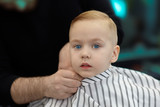 Fototapeta  - Cute blond caucasian baby boy with blue eyes in a barber shop after having haircut by hairdresser. Hands of stylist. Children fashion. Indoors, dark background, copy space.