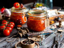 Homemade Canned Hot Tomato Sauce Adjika In Two Glass Jars Standing On Wooden Board