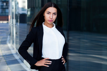 Serious caucasian successful businesswoman in suit, white shirt looking at the camera with a confident look. Brunette stylish glamorous female, girl outdoor, outside office building, business centre 