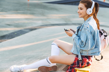 Poster - Image of brunette smiling girl listening music with cellphone and headphones while sitting on sports ground