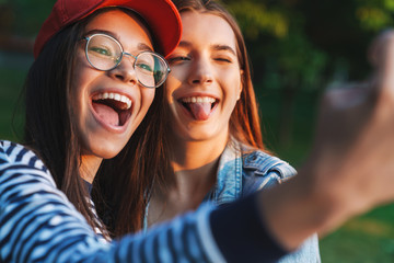 Canvas Print - Image of two laughing girls sticking out her tongue and taking selfie while walking in green park