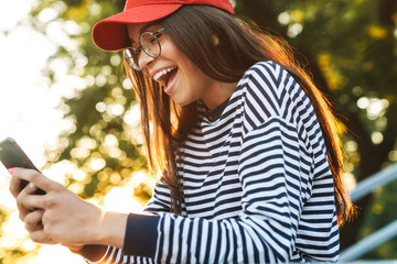 Canvas Print - Image of delighted caucasian girl smiling and using cellphone while walking in green park