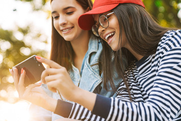 Canvas Print - Image closeup of two happy girls using cellphone and smiling while walking in green park