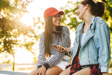 Canvas Print - Image of two laughing girls listening music with earphones on cellphone while sitting on railing in green park