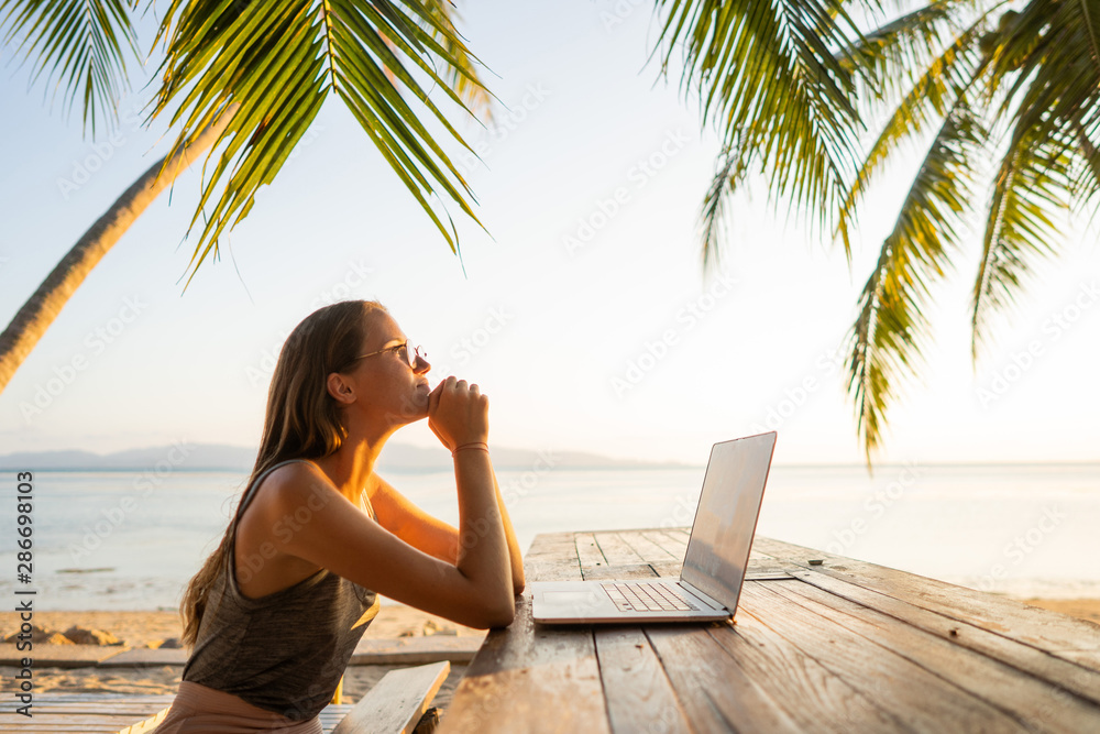 freelancer girl with a computer among tropical palm trees work on the island in sunset - obrazy, fototapety, plakaty 