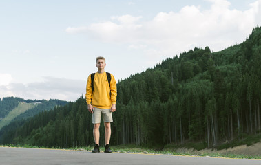 Happy caucasian male hiker tourist standing at the fir forest background in the mountains. Smiling young traveler man looking at the camera, posing for the photo at the woodland landscape.