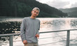 © bodnarphoto - Man in a grey sweatshirt and hat enjoying the view on the lake and fir forest. Young male tourist looking at the camera near the pond in the moutain woodland. Evening landscape.