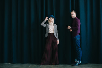 Wall Mural - full length view of stylish young man in glasses and blonde woman in hat near curtain