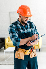 Wall Mural - happy bearded worker in safety helmet writing while holding clipboard