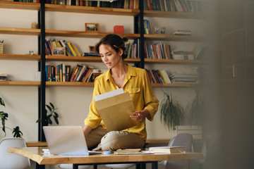 Aufkleber - Young beautiful woman in yellow shirt sitting on desk with laptop and papers while dreamily working in modern office