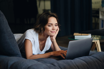 Wall Mural - Young attractive smiling woman lying on sofa happily working on new project with laptop in office