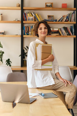 young attractive smiling woman in white shirt happily looking in camera sitting on desk with papers 