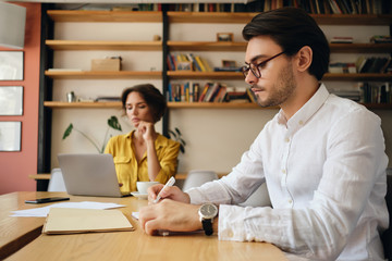 Fototapete - Young attractive man in eyeglasses sitting at the table thoughtfully writing in notepad with colleague on background at work in office