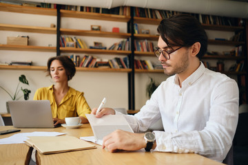 Fototapete - Young handsome man in eyeglasses sitting at the table thoughtfully planning work with colleague on background in office