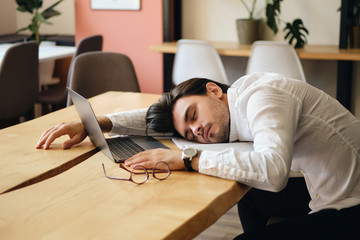 Young exhausted man sitting at the table with laptop while sleeping at work in modern office