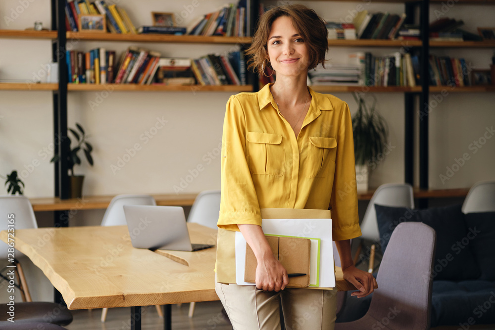 Young beautiful woman in yellow shirt leaning on desk with notepad and papers in hand while happily looking in camera in modern office - obrazy, fototapety, plakaty 