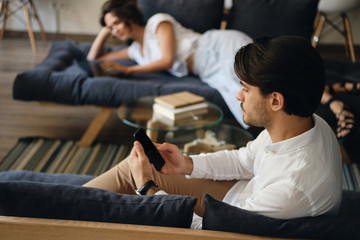 Aufkleber - Side view of young businessman sitting on sofa with colleague on background while thoughtfully using cellphone in co-working space