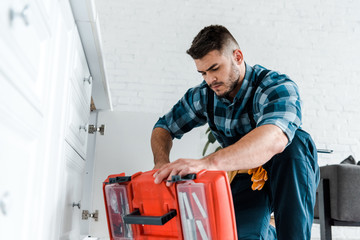 Wall Mural - selective focus of handsome handyman looking at opened toolbox in kitchen