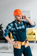 © LIGHTFIELD STUDIOS - handsome worker in safety helmet talking on smartphone while standing with hand on hip