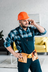 Wall Mural - handsome worker in safety helmet talking on smartphone while standing with hand on hip