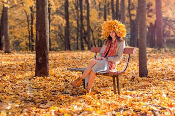 a girl sits in a park on a bench in a wreath of autumn leaves, a golden crown on her head from yellow maple leaves