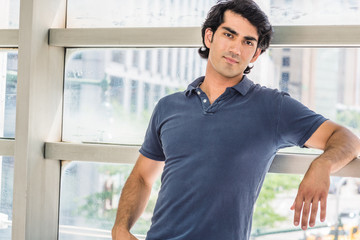 Wall Mural - Portrait of Young Mix Race American College Student in New York City. Young handsome man wearing blue short sleeve shirt, standing against glass wall on school campus, relaxing, thinking..