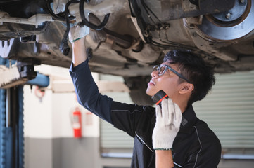 Asian male mechanical hold and shining flashlight to examine car under chassis of automotive vehicle. Safety suspension inspection check service maintenance for customer before road trip concept