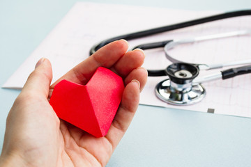 Cardiology and medical concept. Red heart in a hand, a stethoscope and a cardiogram in the background