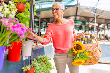 Attractive mature woman shopping in an outdoors fresh flowers market stall, buying and picking from a large variety of colorful floral bouquets during a sunny day in the city.