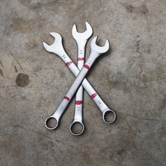 Square image of three chrome wrenches of different sizes lying crossed on concrete floor.