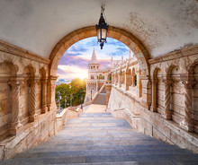 Budapest, Hungary. Ancient Fishermans Bastion Castle. View At Tower From Stairs Under Arch Along Gallery With Columns And Street Lantern. Picturesque Evening Sunset With Clouds On Sky. Famous.