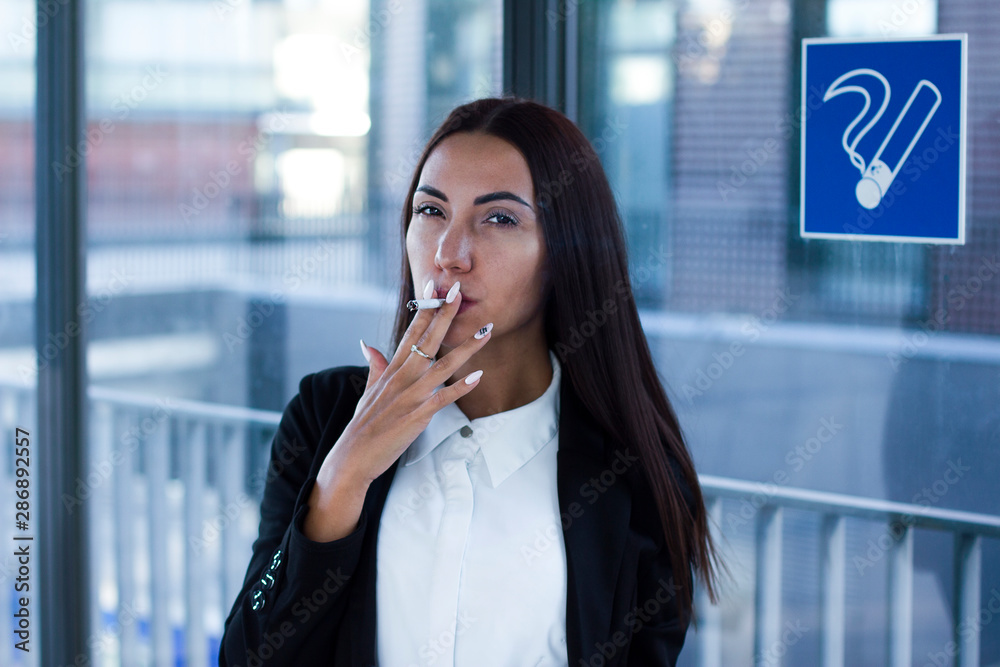 Photo Stock Young stylish glamorous businesswoman puffed by a cigarette,  looking at camera in a smoking room, place next to sign smoking is allowed.  Formal dressed office female worker in smoking area
