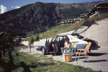 1982 Vintage Analog Camera Image Of A Young Family Driving A Light Blue Saab 900 During Summer Vacation Having A Picnic In The Swiss Alps.