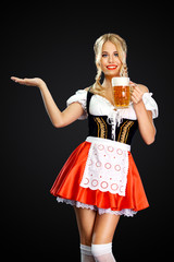 Smiling sexy oktoberfest girl waitress, wearing a traditional Bavarian or german dirndl, serving two big beer mugs with drink isolated on black background. Woman pointing to looking left.