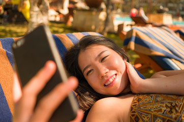 Wall Mural - young beautiful and happy tourist Asian Korean woman taking selfie at tropical resort pool hammock smiling carefree and natural in Summer holidays getaway