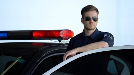 Wall Mural - Serious patrolman in sunglasses monitoring road standing near door of police car