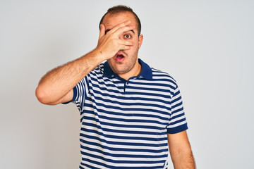 Sticker - Young man wearing casual striped polo standing over isolated white background peeking in shock covering face and eyes with hand, looking through fingers with embarrassed expression.