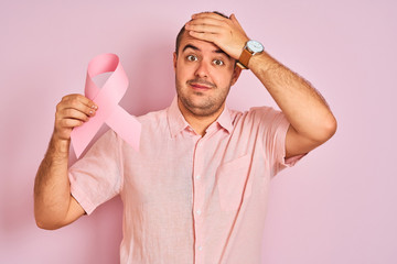 Sticker - Young man holding cancer ribbon symbol standing over isolated pink background stressed with hand on head, shocked with shame and surprise face, angry and frustrated. Fear and upset for mistake.