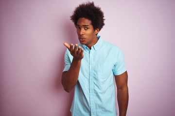 Poster - Young american man with afro hair wearing blue shirt standing over isolated pink background looking at the camera blowing a kiss with hand on air being lovely and sexy. Love expression.