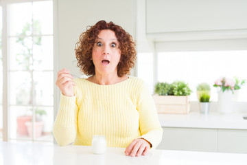 Poster - Senior woman eating a healthy natural yogurt at home scared in shock with a surprise face, afraid and excited with fear expression