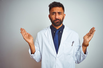 Wall Mural - Young indian doctor man standing over isolated white background clueless and confused expression with arms and hands raised. Doubt concept.