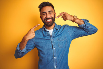 Wall Mural - Young indian man wearing denim shirt standing over isolated yellow background smiling cheerful showing and pointing with fingers teeth and mouth. Dental health concept.