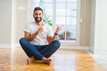 Sticker - Handsome hispanic man wearing casual t-shirt sitting on the floor at home smiling and looking at the camera pointing with two hands and fingers to the side.