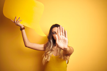 Canvas Print - Young beautiful woman holding speech bubble over yellow isolated background with open hand doing stop sign with serious and confident expression, defense gesture