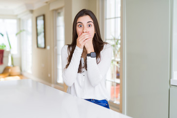 Canvas Print - Beautiful young woman sitting on white table at home shocked covering mouth with hands for mistake. Secret concept.