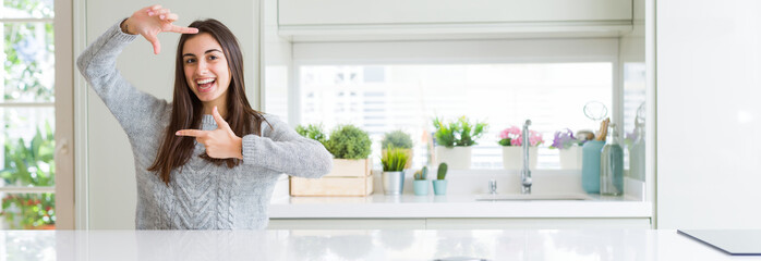 Poster - Wide angle picture of beautiful young woman sitting on white table at home smiling making frame with hands and fingers with happy face. Creativity and photography concept.