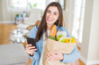 © Krakenimages.com - Young woman holding a paper bag full of fresh groceries and using smartphone app for supermarket delivery
