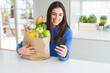 © Krakenimages.com - Young woman holding a paper bag full of fresh groceries and using smartphone app for supermarket delivery